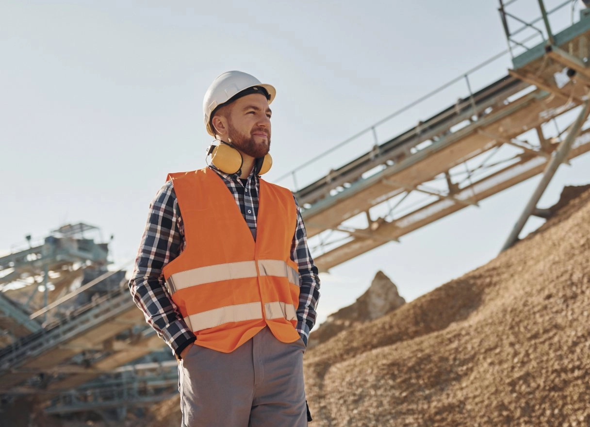 Mine worker in a hard hat and high-visibility vest standing near a conveyor and aggregate pile